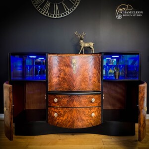 May include: A vintage wooden bar cabinet with a curved front, two doors, and two drawers. The cabinet has a black finish and features a decorative brass accent on the front. The interior of the cabinet is lit with blue LED lights.