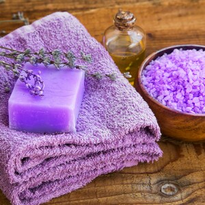 May include: Purple bath salts in a wooden bowl, a purple bar of soap, a purple towel, and a clear glass bottle of oil on a wooden surface.