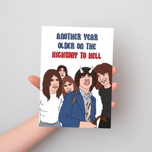 May include: A rectangular greeting card featuring a colorful cartoon illustration of a rock band. The text at the top reads "ANOTHER YEAR OLDER ON THE HIGHWAY TO HELL" in blue and red lettering. The band members are depicted with brown hair.