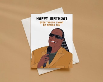 May include: A white greeting card featuring a cartoon illustration of a person wearing dark sunglasses and a tan blazer, holding a black microphone. The text reads HAPPY BIRTHDAY EVEN THOUGH I WONT BE SEEING YOU. The card rests on a white envelope.