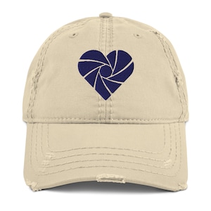 May include: A beige distressed baseball cap featuring an embroidered dark navy blue heart shape composed of camera aperture blades. The cap has a curved brim with frayed edges and a structured front panel with matching stitching throughout the design.