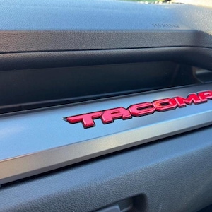 以下が含まれることがあります： 赤い盛り上がった文字で「TACOMA」と書かれた車のダッシュボードのクローズアップ。文字は銀色のトリムピースの中央に配置されています。ダッシュボードは黒とグレーです。