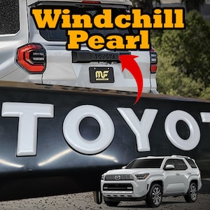 Puede incluir: Un SUV Toyota blanco con el texto "Windchill Pearl" en fuente amarilla y negra. La palabra "TOYOTA" se muestra en letras 3D blancas grandes. Una flecha roja apunta al emblema de Toyota.