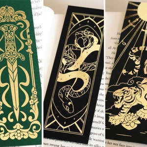 Può includere: Tre design di segnalibri in lamina dorata su sfondi di colori diversi. Il primo segnalibro è verde con un design a spada dorata. Il secondo segnalibro è nero con un design a serpente e rosa dorati. Il terzo segnalibro è nero con un design a tigre dorata.