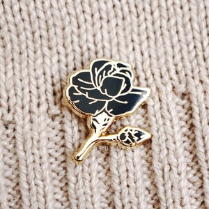 May include: A gold-toned pin featuring a black rose design. The rose is stylised and has a long stem with a bud.