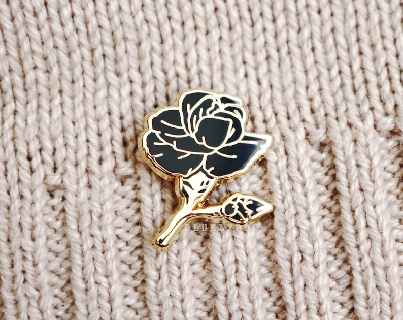 May include: A gold-toned pin featuring a black rose design. The rose is stylised and has a long stem with a bud.
