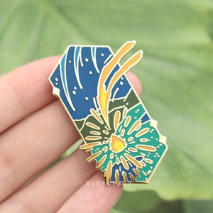 Op de afbeelding: Een gouden emaille pin met een blauw, groen en geel ontwerp. Het ontwerp toont een gestileerd vuurwerk dat explodeert met een geel centrum en een groene en blauwe achtergrond. De pin heeft de vorm van een rechthoek met afgeronde hoeken.