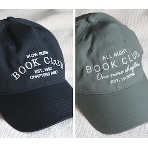 May include: Two baseball caps, one black and one olive green. The black cap has white embroidered text that reads "SLOW BURN BOOK CLUB EST. 1952 CHAPTERS AGO." The green cap has white embroidered text that reads "ALL NIGHT BOOK CLUB One more chapter EST. 11:30PM."