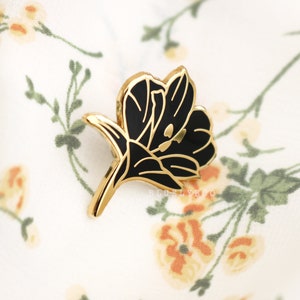 Puede incluir: Pin de flor de tono dorado con detalles en esmalte negro. El pin presenta una flor estilizada con una sola flor y un tallo.