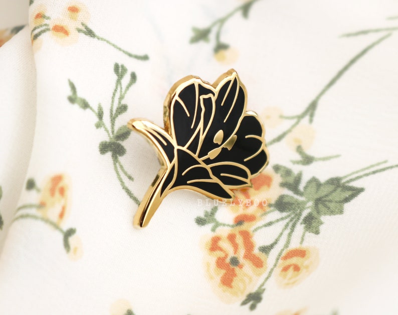 Puede incluir: Pin de flor de tono dorado con detalles en esmalte negro. El pin presenta una flor estilizada con una sola flor y un tallo.