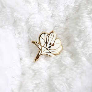 Puede incluir: Un pin de flor de esmalte blanco con detalles dorados. La flor est&aacute; estilizada y tiene un dise&ntilde;o delicado.