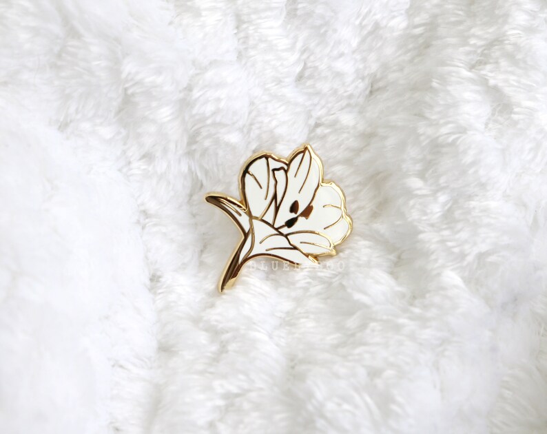 Puede incluir: Un pin de flor de esmalte blanco con detalles dorados. La flor est&aacute; estilizada y tiene un dise&ntilde;o delicado.