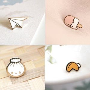May include: Four enamel pins featuring a paper airplane, a popsicle, a dumpling, and a fortune cookie. The pins are gold-toned with white, pink, and orange accents.