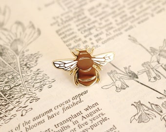 May include: Gold-toned bee pin with white wings. The pin is on a vintage book page with botanical illustrations.