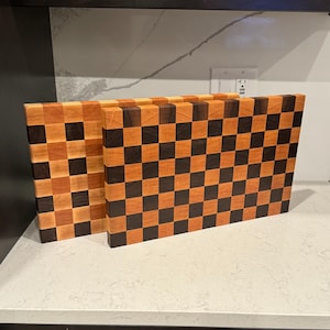 May include: Two end-grain cutting boards with a checkerboard pattern. The boards are made of alternating squares of light and dark wood, creating a visually appealing design. The boards are rectangular and appear to be handmade.