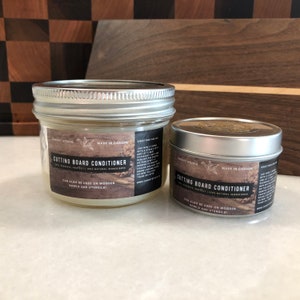Op de afbeelding: Twee potten met snijplank conditioner, een in een glazen pot en een in een metalen blik. Beide potten hebben een wit etiket met zwarte tekst die "Snijplank conditioner" en "Kan ook worden gebruikt voor houten kommen en bestek!" leest. De etiketten bevatten ook de bedrijfsnaam "Canopy Studio" en "Gemaakt in Canada".