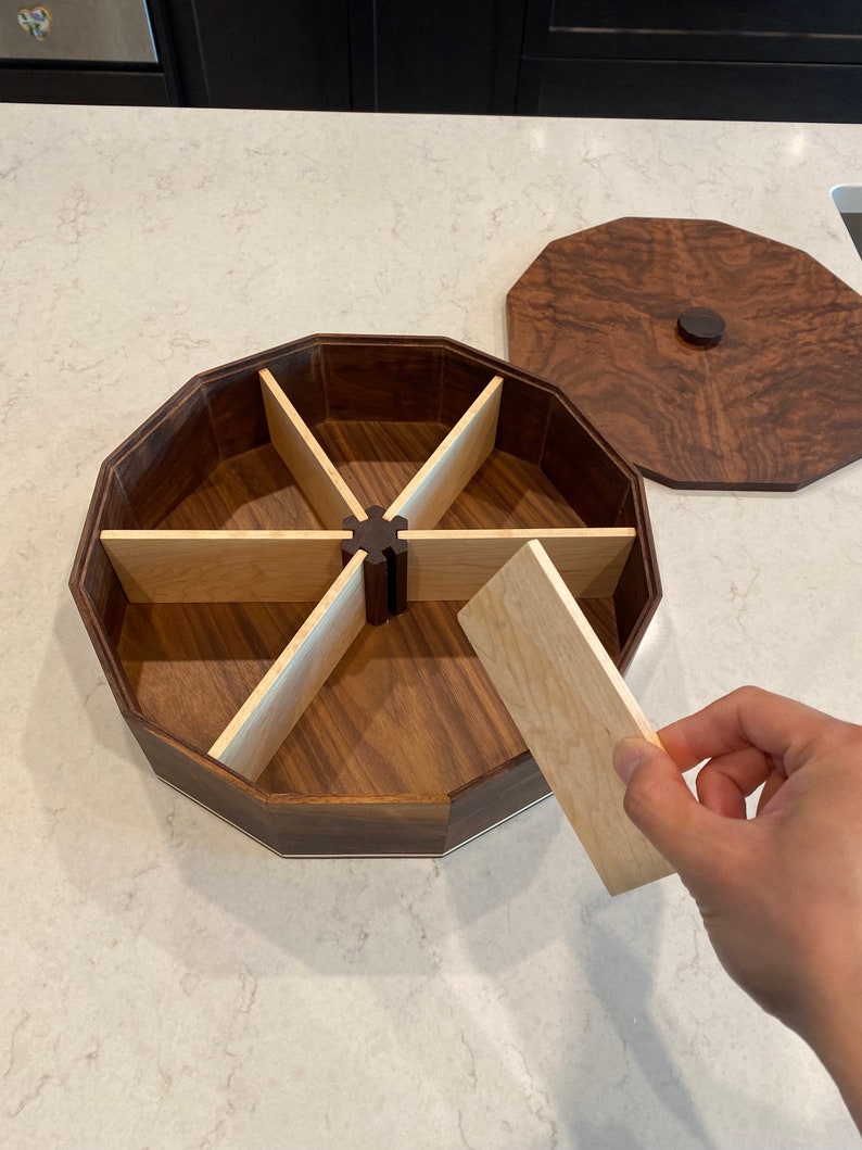 May include: A wooden octagonal box with a lid, divided into six compartments. The box is made of walnut wood with maple wood dividers. The lid is also made of walnut wood and has a small knob on top.