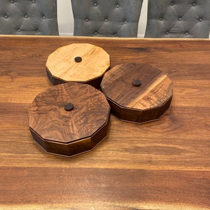 May include: Three wooden octagonal boxes with lids. Two boxes are made of dark wood and one is made of light wood. The boxes are sitting on a wooden table.
