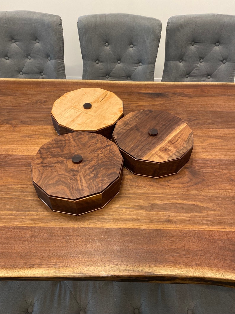 May include: Three wooden octagonal boxes with lids. Two boxes are made of dark wood and one is made of light wood. The boxes are sitting on a wooden table.