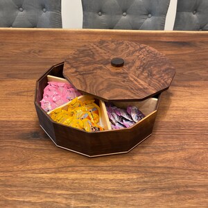 May include: A wooden octagonal box with a lid, divided into six compartments. The box is filled with small packages of candy in pink, yellow, and purple.