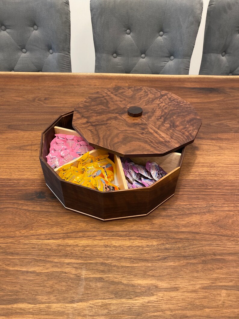 May include: A wooden octagonal box with a lid, divided into six compartments. The box is filled with small packages of candy in pink, yellow, and purple.