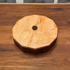 May include: A wooden box with a light wood top and a dark wood base. The box has a round shape with a slightly octagonal design. A small, round, dark knob is in the center of the top.