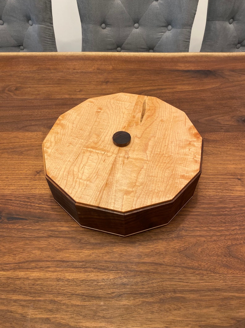 May include: A wooden box with a light wood top and a dark wood base. The box has a round shape with a slightly octagonal design. A small, round, dark knob is in the center of the top.