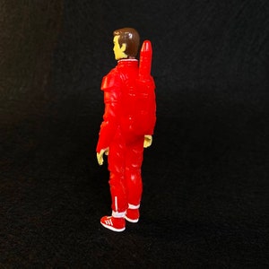 K&ouml;nnte beinhalten: Eine rote Actionfigur eines Mannes in einem roten Overall mit einem gelben Jetpack auf dem R&uuml;cken. Die Figur hat braunes Haar und tr&auml;gt wei&szlig;e Turnschuhe.