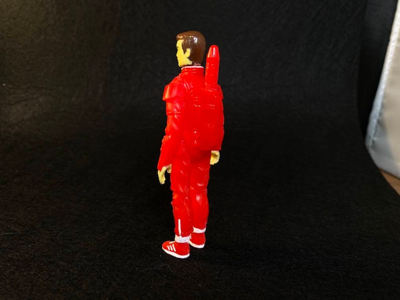 K&ouml;nnte beinhalten: Eine rote Actionfigur eines Mannes in einem roten Overall mit einem gelben Jetpack auf dem R&uuml;cken. Die Figur hat braunes Haar und tr&auml;gt wei&szlig;e Turnschuhe.