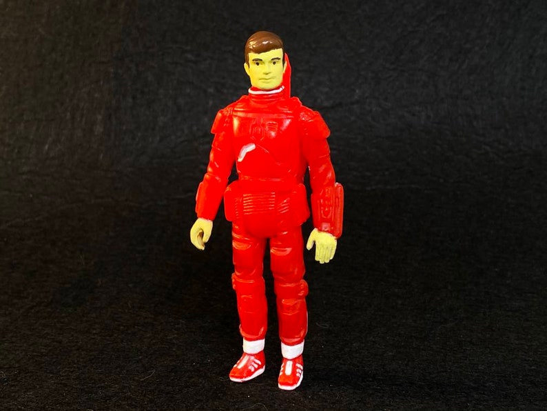 K&ouml;nnte beinhalten: Eine rote Plastik-Actionfigur einer Person, die einen roten Raumanzug mit gelbem Gesicht und braunen Haaren tr&auml;gt. Die Figur steht mit den Armen an den Seiten und tr&auml;gt rote Schuhe mit wei&szlig;en Akzenten.