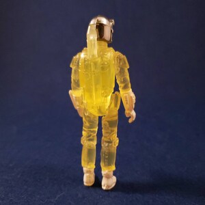 May include: A yellow translucent action figure of a bounty hunter with a silver helmet and a jetpack. The figure is standing on a blue surface.