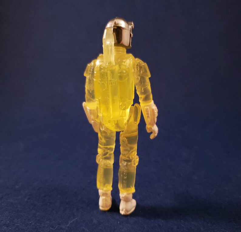 May include: A yellow translucent action figure of a bounty hunter with a silver helmet and a jetpack. The figure is standing on a blue surface.