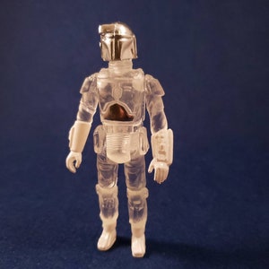 K&ouml;nnte beinhalten: Eine durchsichtige Plastik-Actionfigur des Star Wars-Charakters Boba Fett mit silbernem Helm und R&uuml;stung. Die Figur steht mit den Armen an den Seiten.
