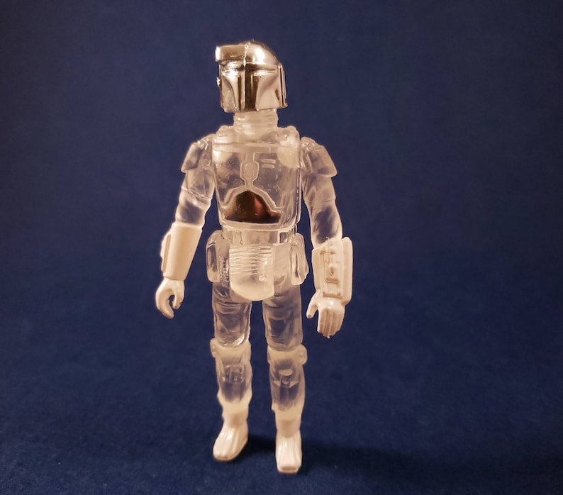 K&ouml;nnte beinhalten: Eine durchsichtige Plastik-Actionfigur des Star Wars-Charakters Boba Fett mit silbernem Helm und R&uuml;stung. Die Figur steht mit den Armen an den Seiten.