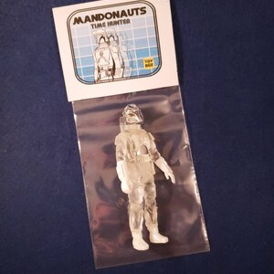 K&ouml;nnte beinhalten: Eine transparente Plastik-Actionfigur eines Mandalorianer-Charakters, verpackt in einer Plastikt&uuml;te mit einer wei&szlig;en Karte mit dem Text "Mandonaunts Time Hunter" und einem gelben "Toy Biz"-Logo.