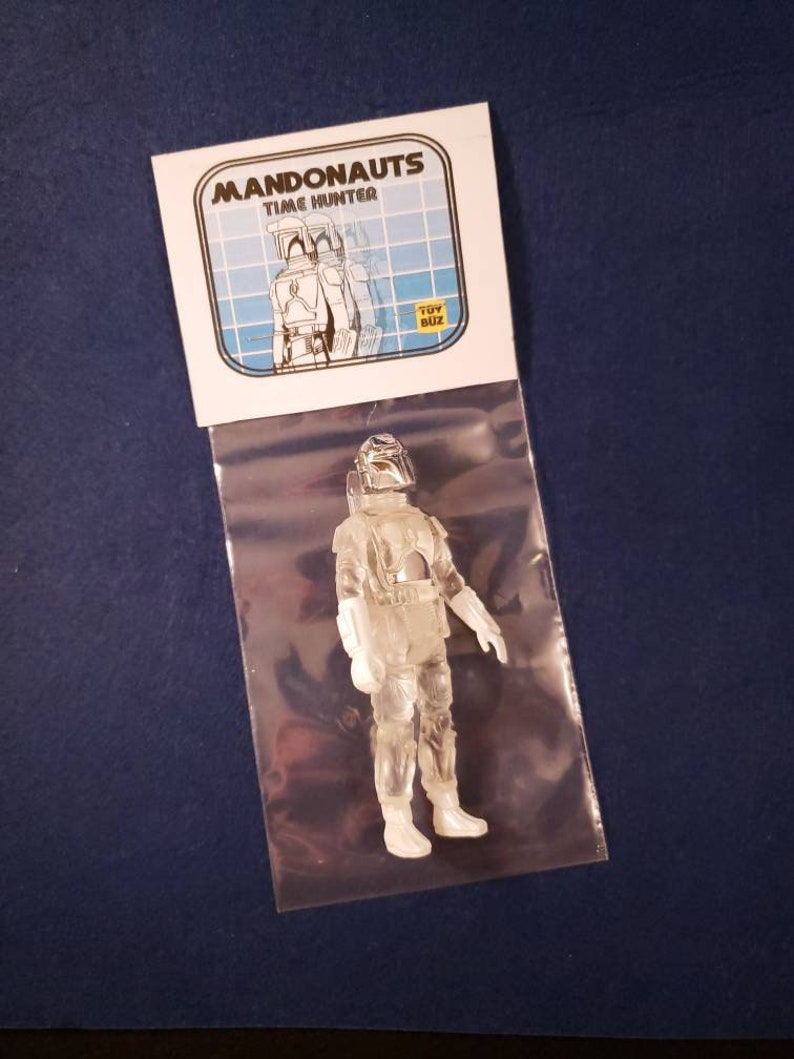 K&ouml;nnte beinhalten: Eine transparente Plastik-Actionfigur eines Mandalorianer-Charakters, verpackt in einer Plastikt&uuml;te mit einer wei&szlig;en Karte mit dem Text "Mandonaunts Time Hunter" und einem gelben "Toy Biz"-Logo.