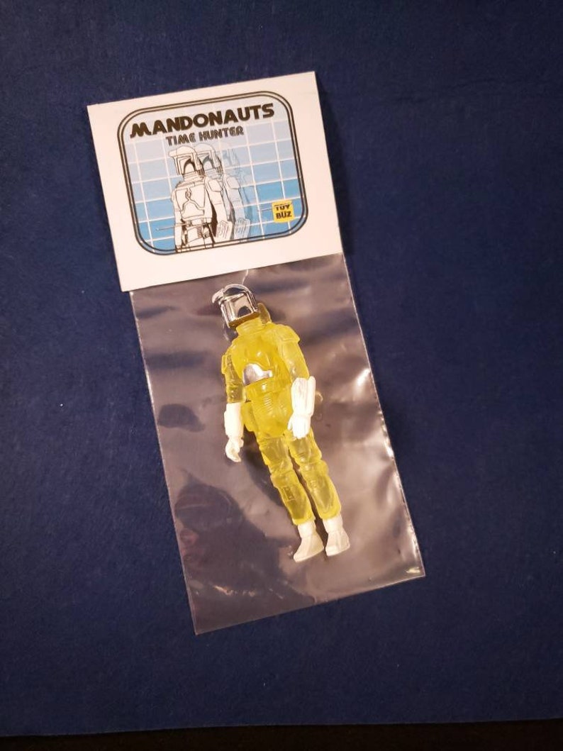 May include: A yellow translucent action figure of a Mandalorian with a white helmet and white armor. The figure is packaged in a clear plastic bag with a white card that reads "Mandonaunts Time Hunter" and "Toy Biz".