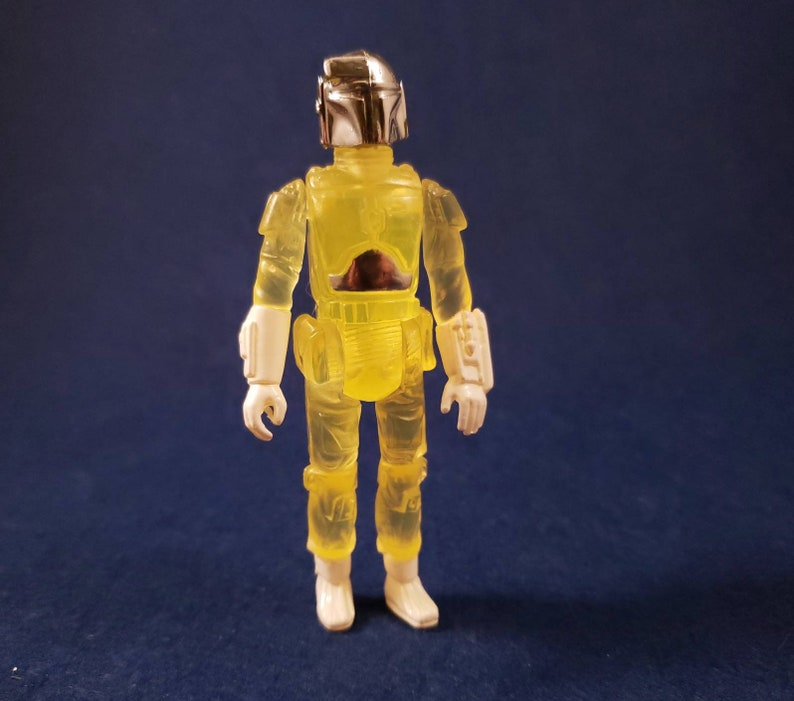 May include: A yellow translucent action figure of a Star Wars character wearing a helmet and armor. The figure has white boots and is standing on a blue background.