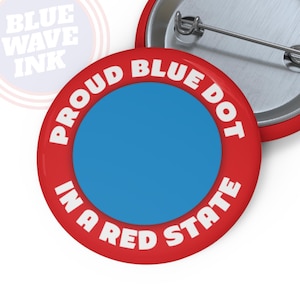 May include: Red and white button with the text "Proud Blue Dot in a Red State". The button has a blue circle in the center.