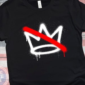 May include: Black t-shirt featuring a white crown design with a red slash through it, created with a spray paint effect. The shirt has rolled-up sleeves and a crew neck. The words "BLUE WAVE INK" are visible on the bottom left.