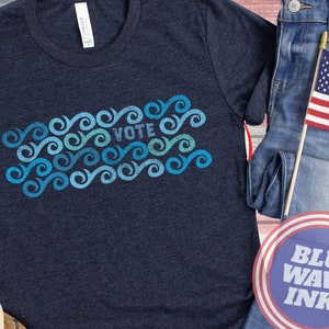 May include: A navy blue t-shirt with a blue and green wave design and the word "VOTE" in white text. The t-shirt is on a wooden surface with a pair of blue jeans and a small American flag.