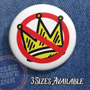 May include: A white button with a yellow crown design crossed out by a red circle and line. The button has the text "3 Sizes Available" in a serif font. The button is on a blue denim background.