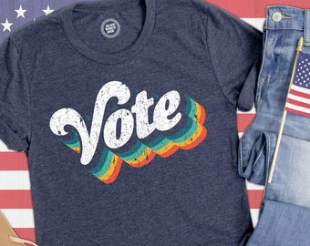 May include: A dark heather navy blue short-sleeve t-shirt featuring the word Vote in a distressed white font. Below the text are three wavy, layered stripes in teal, yellow, and orange. A circular logo on the inner neck reads Blue Wave Ink.