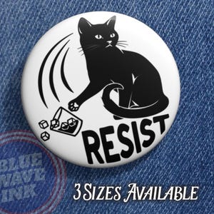 May include: A white button with a black cat illustration knocking over a glass of ice cubes. The word "RESIST" is printed in bold black letters. The text "3 SIZES AVAILABLE" is also visible.