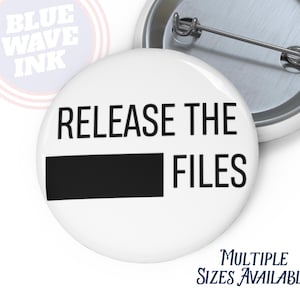 May include: White button with the text "RELEASE THE FILES" in black, with a black rectangle. The button has a metal pin back. The text "MULTIPLE SIZES AVAILABLE" is in blue. A second button is partially visible.