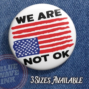 May include: A white button with a distressed American flag design. The button features the text "WE ARE NOT OK" in black, with the flag's stars and stripes in blue and red. The text "3 SIZES AVAILABLE" is also present.
