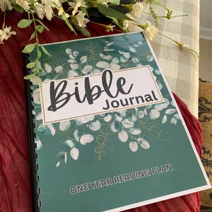 May include: A spiral-bound Bible Journal with a green cover featuring a white and gold eucalyptus leaf design. The title "Bible Journal" is displayed in a white rectangle with a black border. The text "ONE YEAR READING PLAN" is at the bottom.