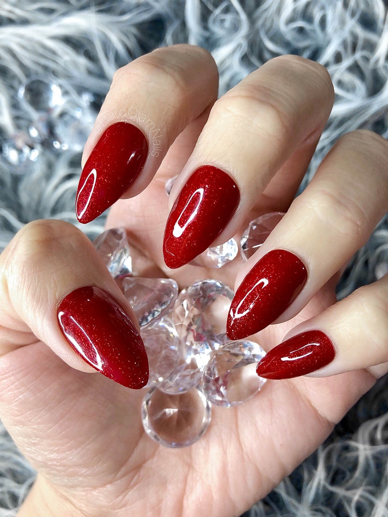 May include: Close-up of a hand with red glitter nail polish on all five nails. The nails are long and pointed, and the glitter is very fine. The hand is holding a pile of clear crystals.
