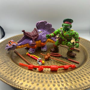 May include: A green turtle action figure wearing a military cap and camouflage outfit, accompanied by a purple dinosaur figure with a brown saddle. Several small plastic accessories are included, such as a red bow, a staff, and a weapon.
