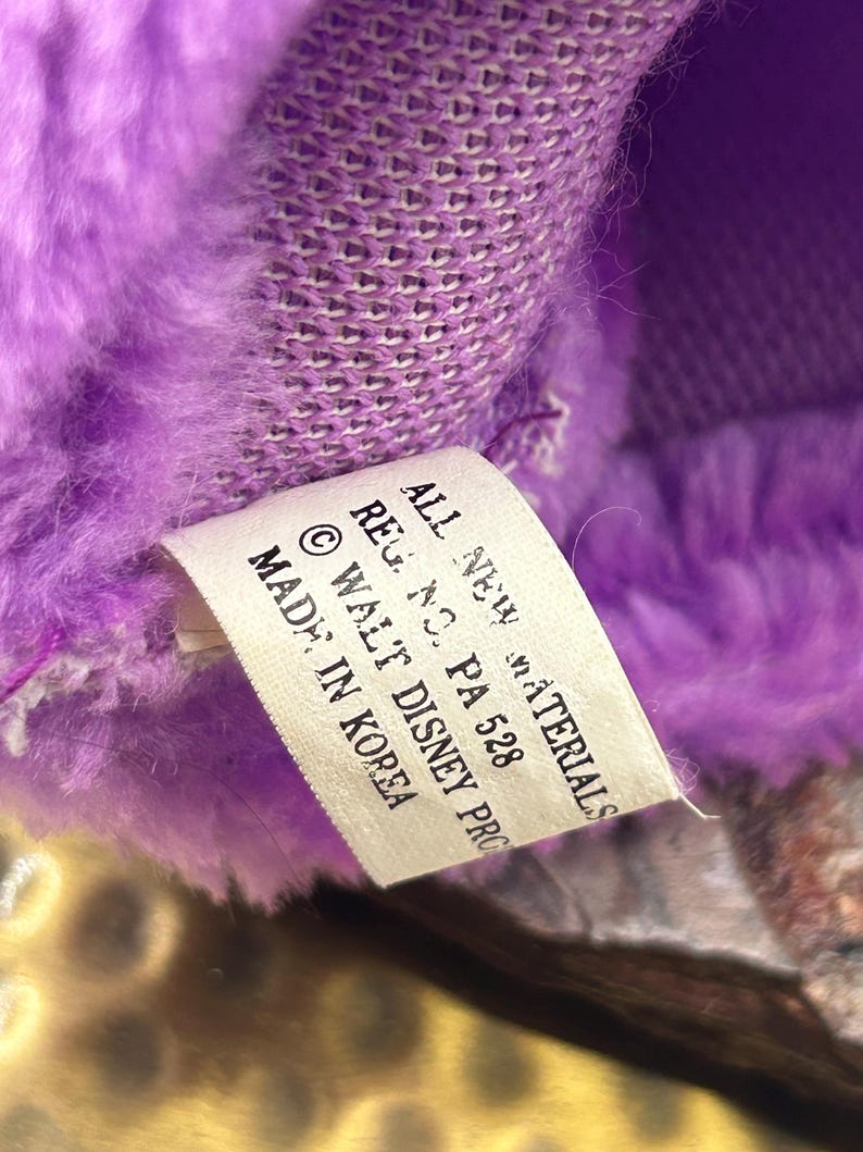 May include: Close-up of a plush toy with purple fur and a tag. The tag reads "ALL NEW MATERIALS, REG. NO. PA 528, &copy; WALT DISNEY PRO, MADE IN KOREA."